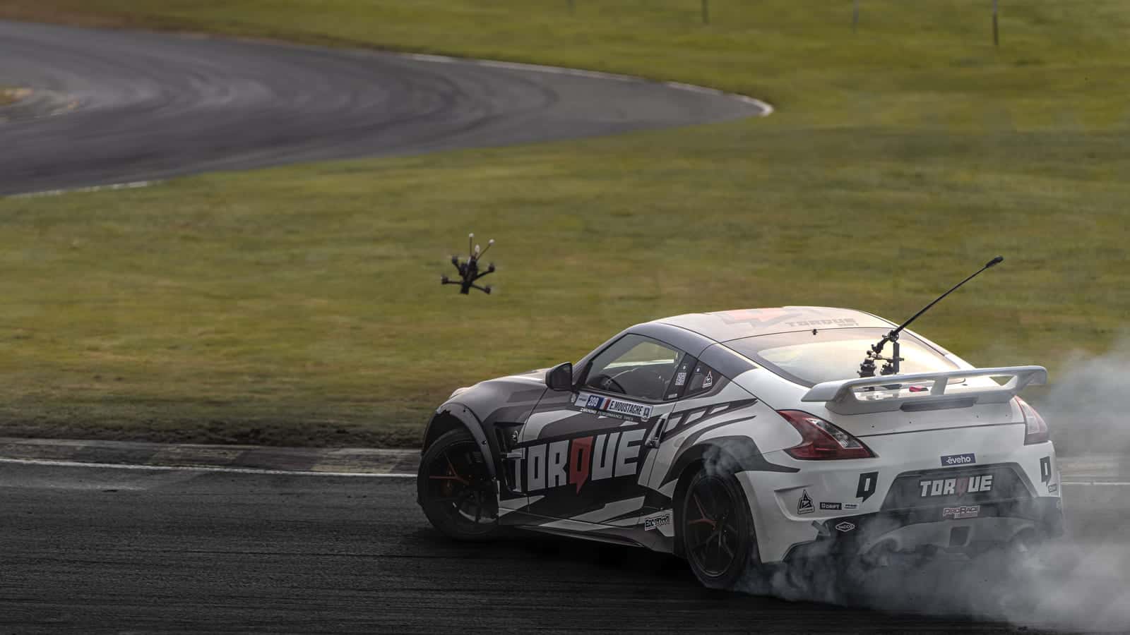 A white drift car trailing tyre smoke on a track while a small FPV drone follows behind it