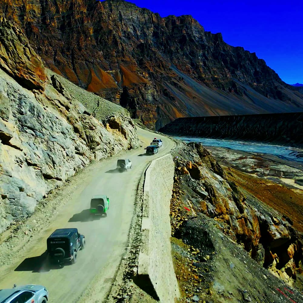 Aerial view of an off-road convoy on a dusty cliff-edge road above a river valley