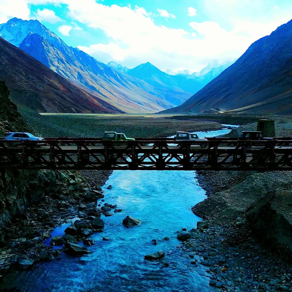 Aerial view of vehicles crossing a steel bridge over a blue river, with snow-capped mountains beyond