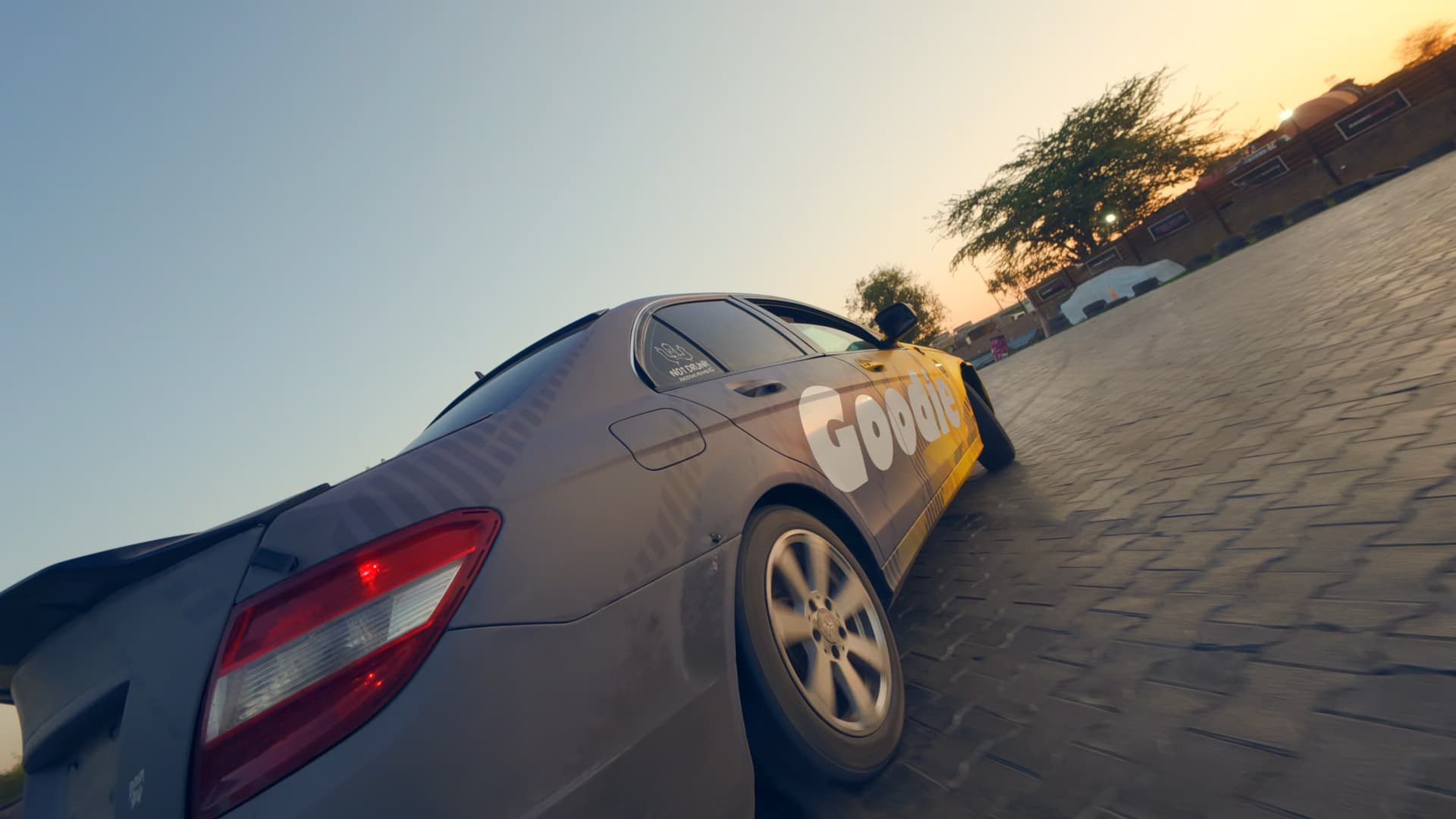 Low-angle view of a grey and yellow sedan with Goodie branding cornering on a paved circuit at sunset, its alloy wheel blurred by motion