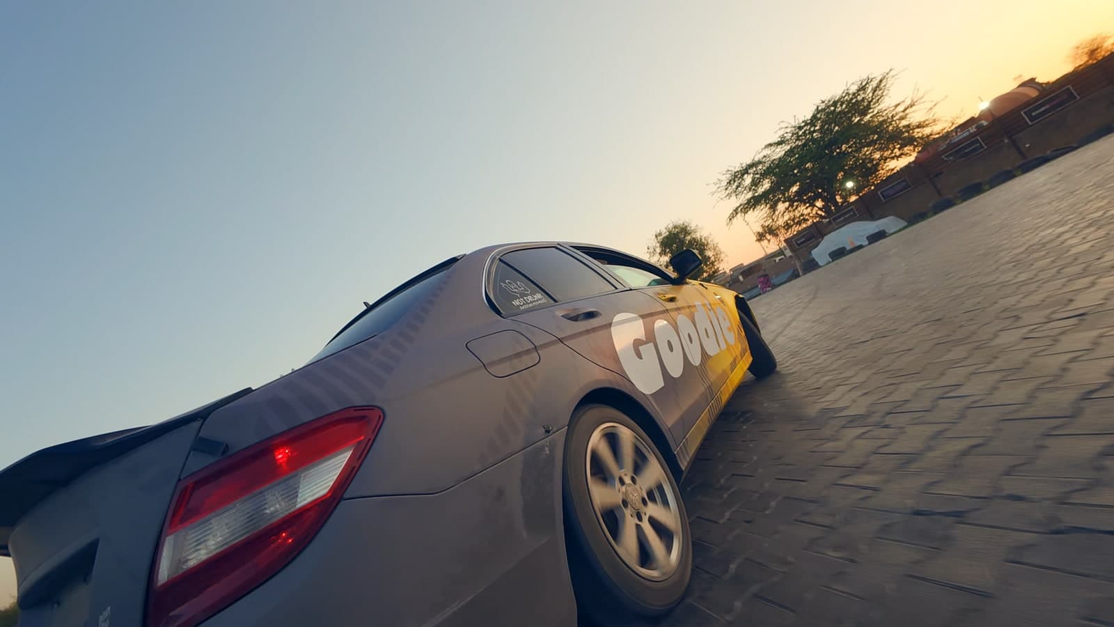 Low-angle view of a grey and yellow sedan with Goodie branding cornering on a paved circuit at sunset, its alloy wheel blurred by motion