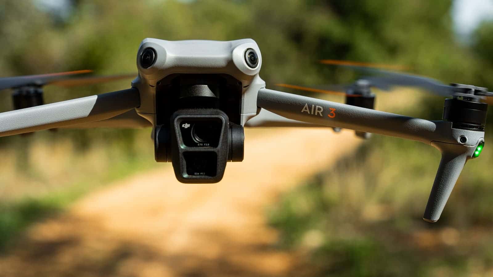 A DJI Air 3 camera drone hovering over a dirt track in a forest, its dual-lens gimbal camera facing the viewer