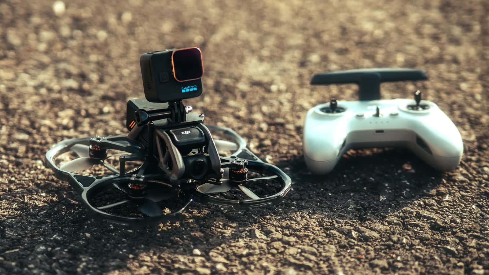 A small FPV drone with a camera on top, resting beside its white controller on gravel