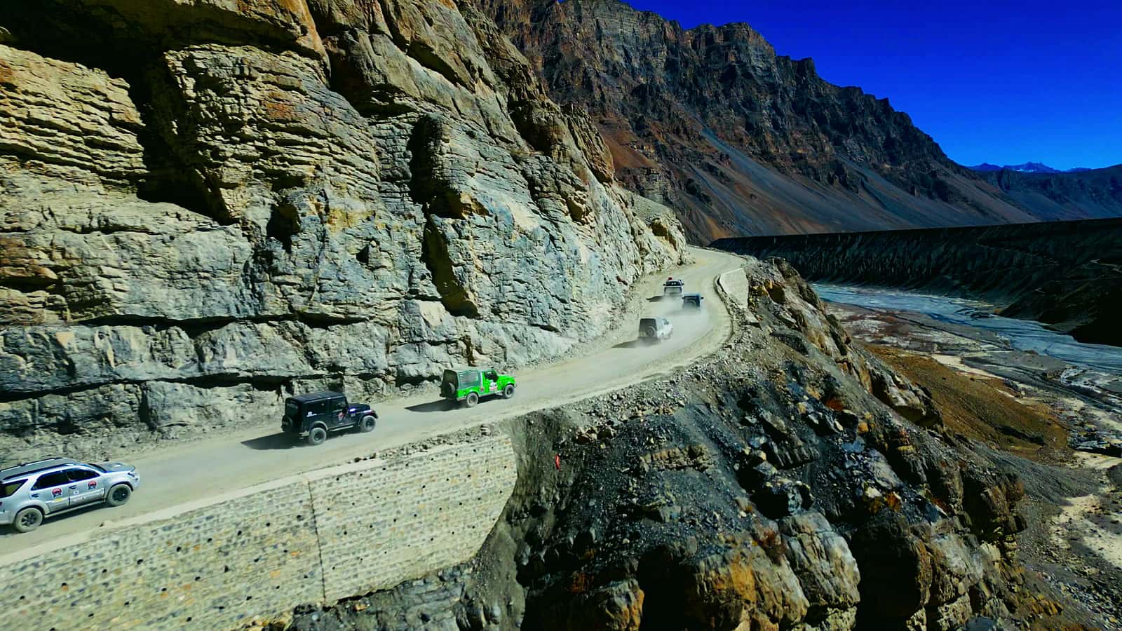 A convoy of off-road vehicles driving along a dusty cliff-edge road above a river valley under a deep blue sky